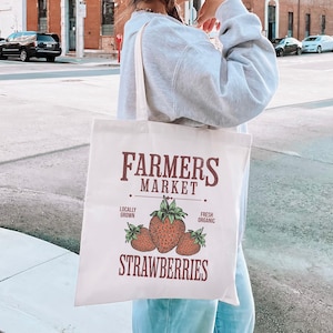 Può includere: Una borsa di tela bianca con un design Farmers Market. Il design presenta le parole "Farmers Market" in un carattere rosso scuro, con la parola "Strawberries" sotto. Tre fragole sono al centro, con le parole "Locally Grown" e "Fresh Organic" su entrambi i lati.