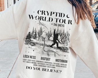 May include: A white sweatshirt with a black graphic print of a Bigfoot-like creature in a forest setting. The text "CRYPTID WORLD TOUR + THE GREYS" is printed above the graphic. Below the graphic, the text "LOCH NESS BIGFOOT MOTHMAN" is printed, followed by a list of cryptids and the question "DO YOU BELIEVE?"