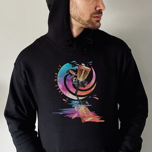 May include: Black hoodie with a front pocket and a drawstring hood. The graphic on the front features a colorful disc golf basket design with the text "JUST RIP IT". The design includes splashes of color and a stylized wave.