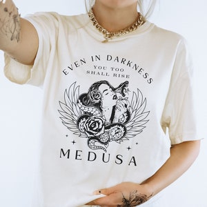 May include: Cream-colored t-shirt featuring a black and white graphic of Medusa with snakes, roses, and wings. The text "EVEN IN DARKNESS YOU TOO SHALL RISE" and "MEDUSA" are printed above and below the graphic. The shirt has short sleeves.