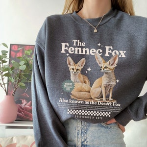 May include: A dark gray sweatshirt with a graphic of two fennec foxes. The text "The Fennec Fox" is printed in a cream color above the foxes. The text "Worlds Smallest Fox" is printed in a green circle below the foxes. The text "Also known as the Desert Fox" is printed below the foxes. The text "Suspiciously Cute" is printed below the desert fox text.