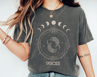 Pode incluir: Uma camiseta cinza com um design gráfico de um círculo com um sol, fases da lua e dois peixes. O texto "Pisces" está abaixo do círculo.