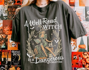 May include: A black t-shirt featuring a graphic print of a witch reading a book. The text on the shirt reads "A Well-Read Witch is a Dangerous Creature".