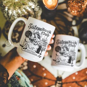 May include: Two white ceramic mugs with black and white illustrations of a witch reading a book. The text on the mugs reads "Salem Book Club" and "Meets Weekly, All Witches Welcome".