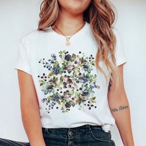 May include: White t-shirt featuring a watercolor design of blackberries, blueberries, and leaves. The design is centered on the shirt and includes shades of green, blue, and purple. The shirt has short sleeves and a crew neck.