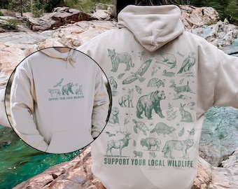 Puede incluir: Una sudadera con capucha beige con un estampado verde de varios animales y el texto "Support Your Local Wildlife". La sudadera tiene capucha y un bolsillo canguro.