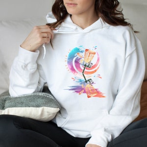 May include: White hoodie featuring a colorful graphic of a disc golf basket and disc with the text "Just Rip It". The design includes shades of blue, purple, orange, and yellow. The hoodie has a drawstring hood and a front pocket.
