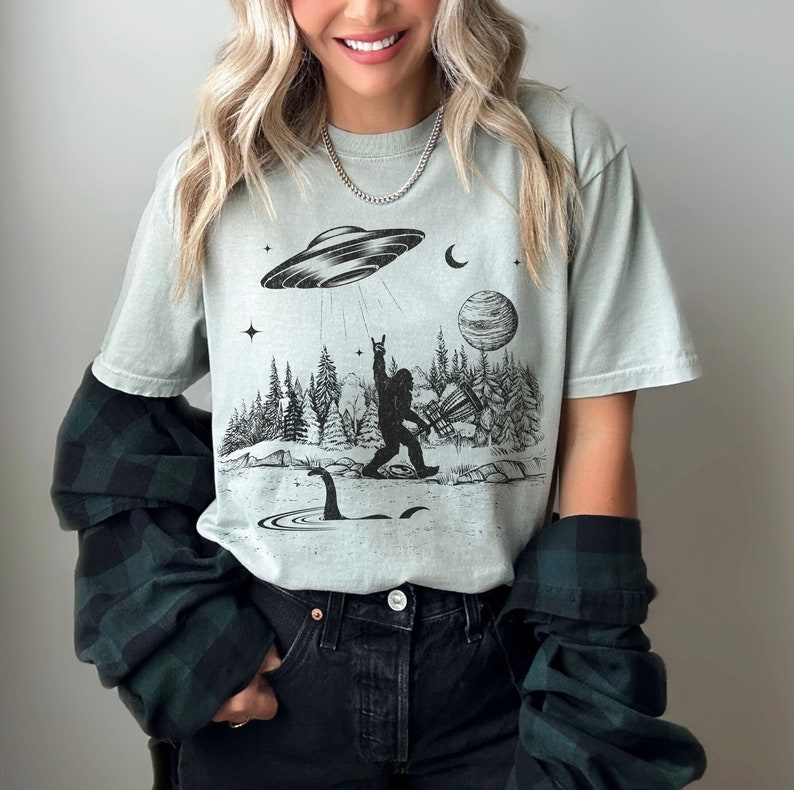 May include: A light green t-shirt featuring a black and white graphic print of a Bigfoot character holding a disc golf disc. The graphic also includes a UFO, a monster, and trees. The shirt has a crew neck and short sleeves.
