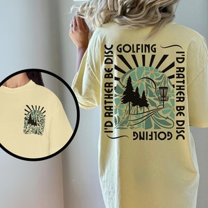 May include: A yellow t-shirt with a graphic design on the back. The design features a disc golf basket, trees, and the text "Golfing I'd rather be disc golfing".
