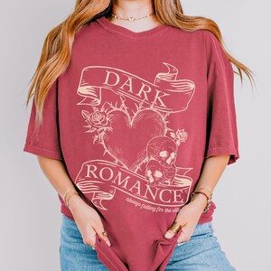 Op de afbeelding: Een maroon t-shirt met een grafisch ontwerp van een hart met doornen, twee schedels en een banner met de tekst "DARK ROMANCE always falling for the villain".