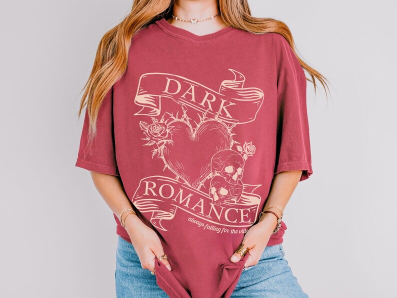 Op de afbeelding: Een maroon t-shirt met een grafisch ontwerp van een hart met doornen, twee schedels en een banner met de tekst "DARK ROMANCE always falling for the villain".