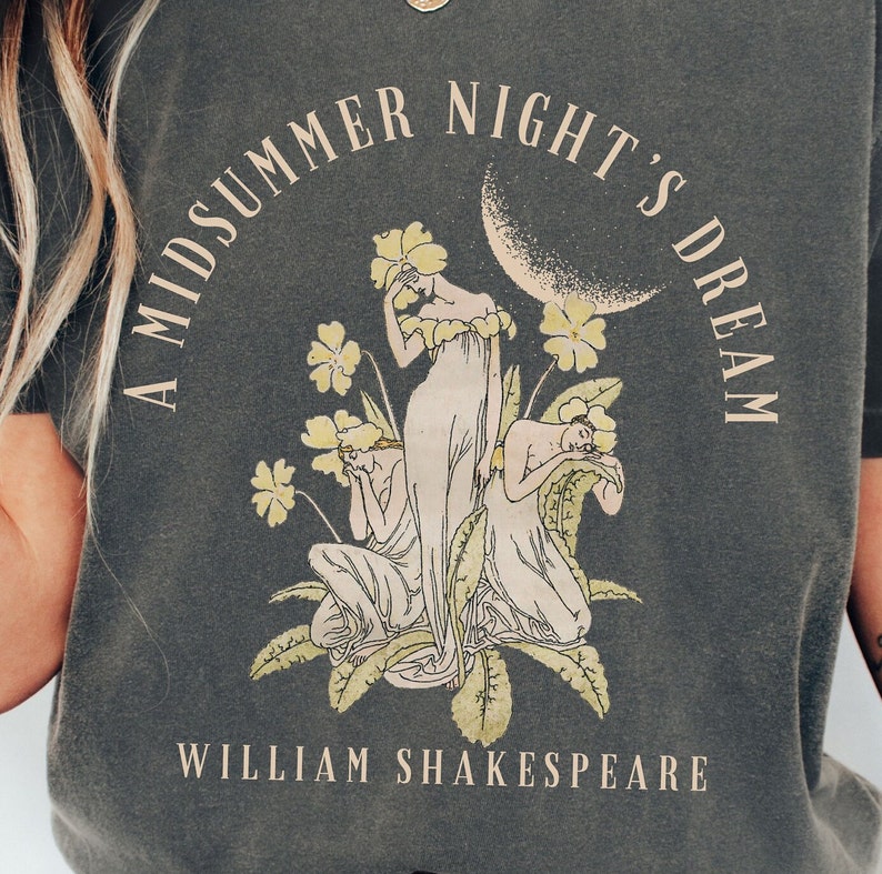 May include: A dark grey t-shirt featuring a vintage-style illustration of three women in a woodland setting. The text "A Midsummer Night's Dream" is printed in a circular arc above the illustration, and "William Shakespeare" is printed below. The illustration includes flowers and a crescent moon.
