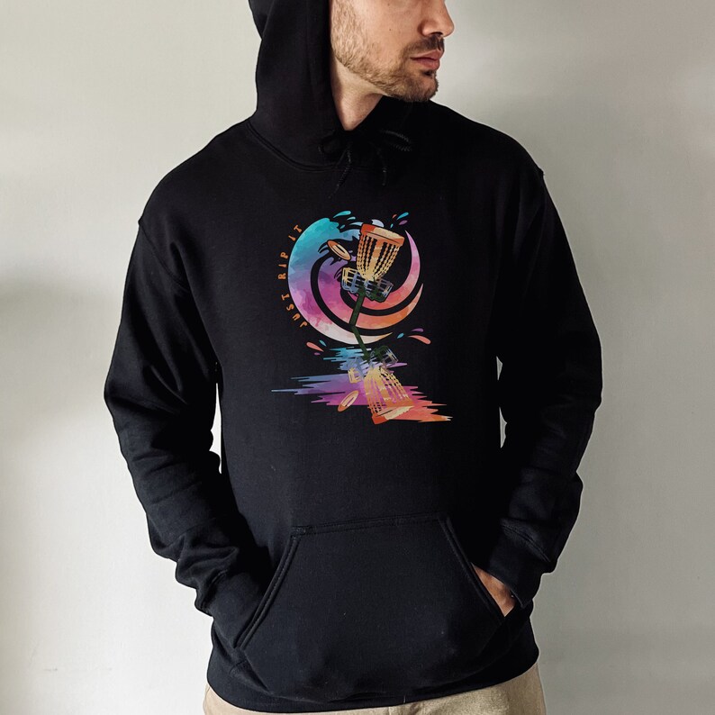 May include: Black hoodie with a front pocket and a drawstring hood. The graphic on the front features a colorful disc golf basket design with the text "JUST RIP IT". The design includes splashes of color and a stylized wave.