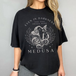 May include: Black t-shirt featuring a Medusa graphic with the text "EVEN IN DARKNESS YOU TOO SHALL RISE". The design includes a Medusa head, snakes, roses, and wings. The shirt is a relaxed fit.