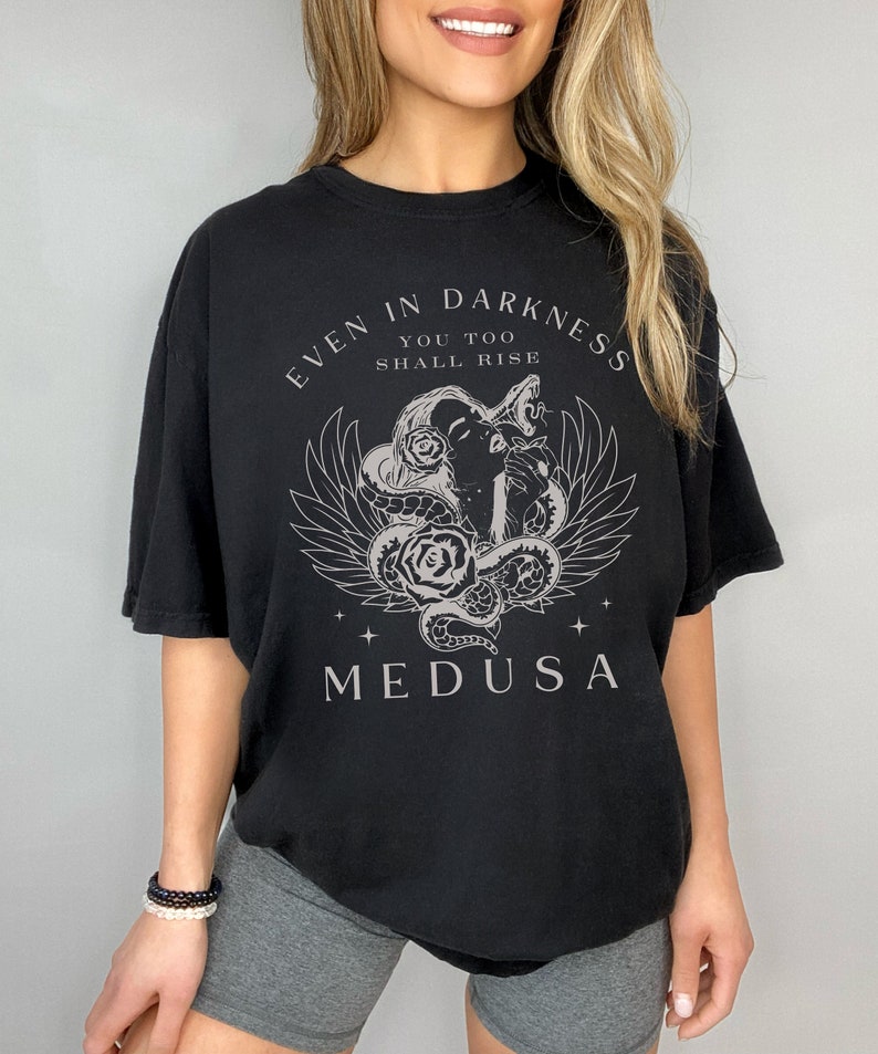 May include: Black t-shirt featuring a Medusa graphic with the text "EVEN IN DARKNESS YOU TOO SHALL RISE". The design includes a Medusa head, snakes, roses, and wings. The shirt is a relaxed fit.