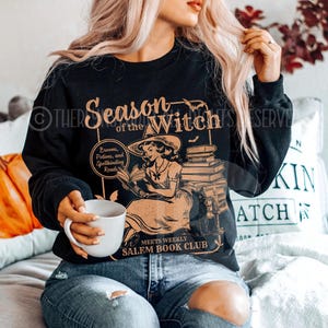May include: Black sweatshirt with a graphic print of a witch reading a book. The text reads "Season of the Witch" and "Meets Weekly Salem Book Club".