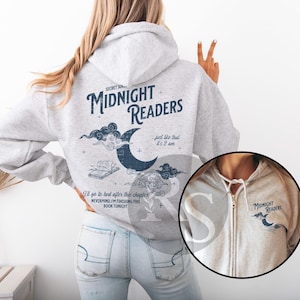 Pu&ograve; includere: Una felpa con cappuccio grigio chiaro con la scritta "MIDNIGHT READERS" e una grafica a mezzaluna. La parte posteriore della felpa ha testo aggiuntivo e una grafica di un libro e nuvole. La parte anteriore ha un piccolo logo.
