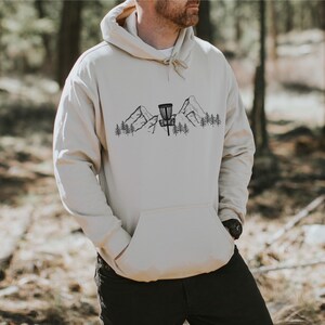 K&ouml;nnte beinhalten: Ein beiger Hoodie mit einer schwarzen Grafik eines Disc-Golf-Korbs vor einer Bergkette mit B&auml;umen.
