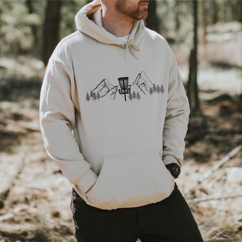 K&ouml;nnte beinhalten: Ein beiger Hoodie mit einer schwarzen Grafik eines Disc-Golf-Korbs vor einer Bergkette mit B&auml;umen.