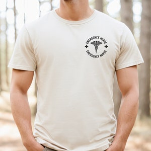 May include: A beige t-shirt with a black graphic of a medical symbol and the text "Emergency Nurse" around it.