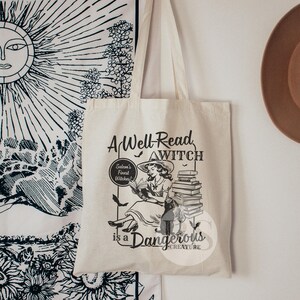K&ouml;nnte beinhalten: Eine wei&szlig;e Canvas-Tragetasche mit einer Schwarzwei&szlig;-Illustration einer Frau, die einen Stapel B&uuml;cher liest. Der Text auf der Tasche lautet "A Well-Read Witch is a Dangerous Creature."