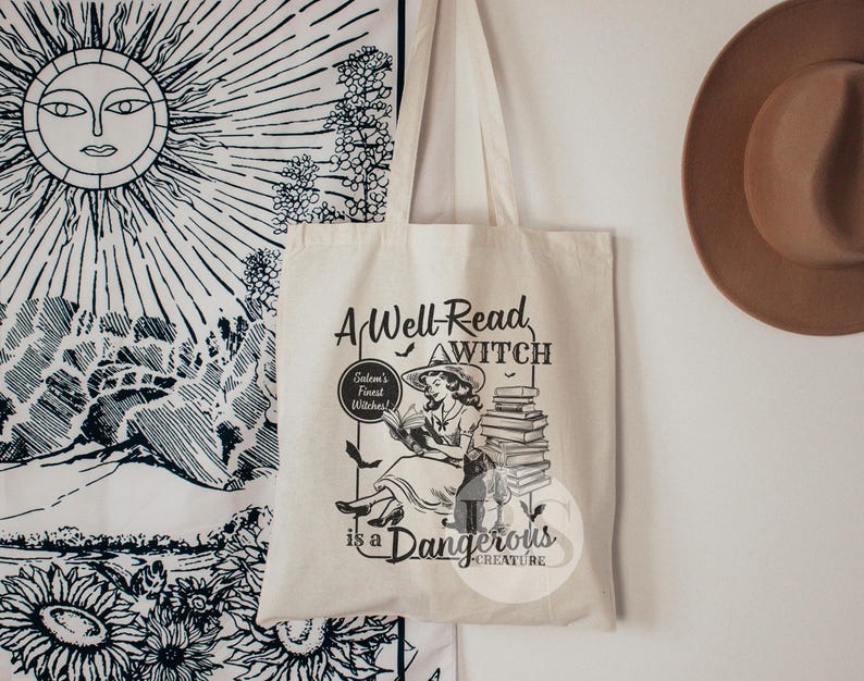 K&ouml;nnte beinhalten: Eine wei&szlig;e Canvas-Tragetasche mit einer Schwarzwei&szlig;-Illustration einer Frau, die einen Stapel B&uuml;cher liest. Der Text auf der Tasche lautet "A Well-Read Witch is a Dangerous Creature."