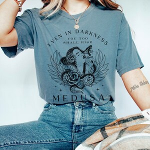 May include: A blue-grey t-shirt featuring a graphic of Medusa with snakes, roses, and wings. The text "EVEN IN DARKNESS YOU TOO SHALL RISE" and "MEDUSA" are printed on the front. The shirt is paired with blue jeans.