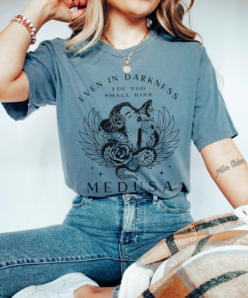 May include: A blue-grey t-shirt featuring a graphic of Medusa with snakes, roses, and wings. The text "EVEN IN DARKNESS YOU TOO SHALL RISE" and "MEDUSA" are printed on the front. The shirt is paired with blue jeans.