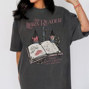 Pu&ograve; includere: Una maglietta grigia oversize con un design grafico di un libro, una bilancia e il testo "The Libra Reader: Finding Beauty & Balance on Every Page".