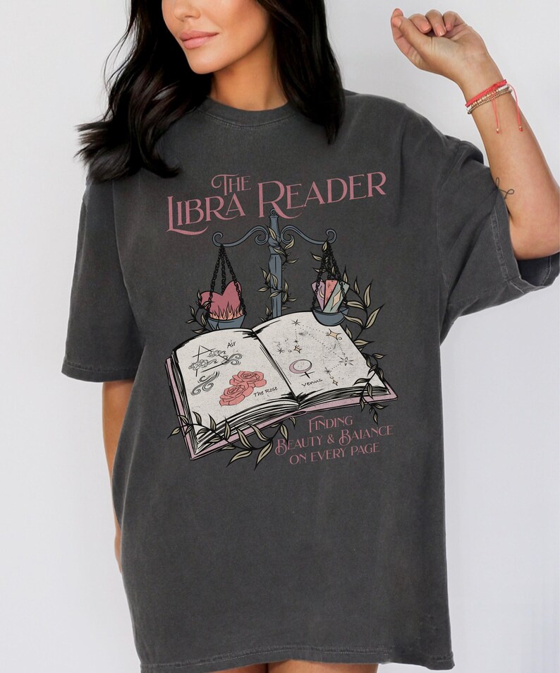 Pu&ograve; includere: Una maglietta grigia oversize con un design grafico di un libro, una bilancia e il testo "The Libra Reader: Finding Beauty & Balance on Every Page".