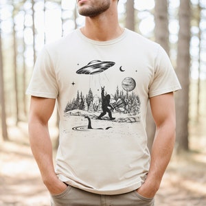 May include: A beige t-shirt featuring a black and white graphic print of a Bigfoot playing a guitar with a UFO above him. The graphic also includes a Loch Ness Monster in the water below.