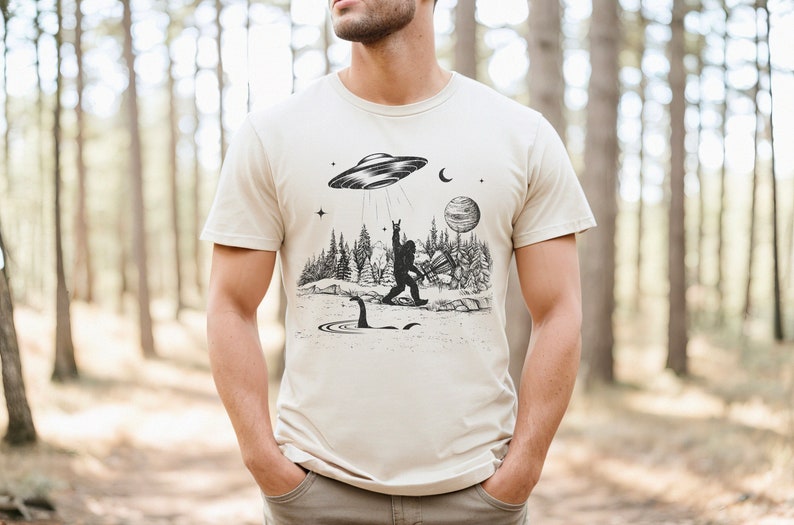 May include: A beige t-shirt featuring a black and white graphic print of a Bigfoot playing a guitar with a UFO above him. The graphic also includes a Loch Ness Monster in the water below.