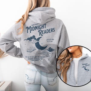 Pu&ograve; includere: Felpa con cappuccio grigia con la scritta "MIDNIGHT READERS" e illustrazioni di una luna crescente, nuvole e un libro. La felpa ha una cerniera e un piccolo logo sul petto.