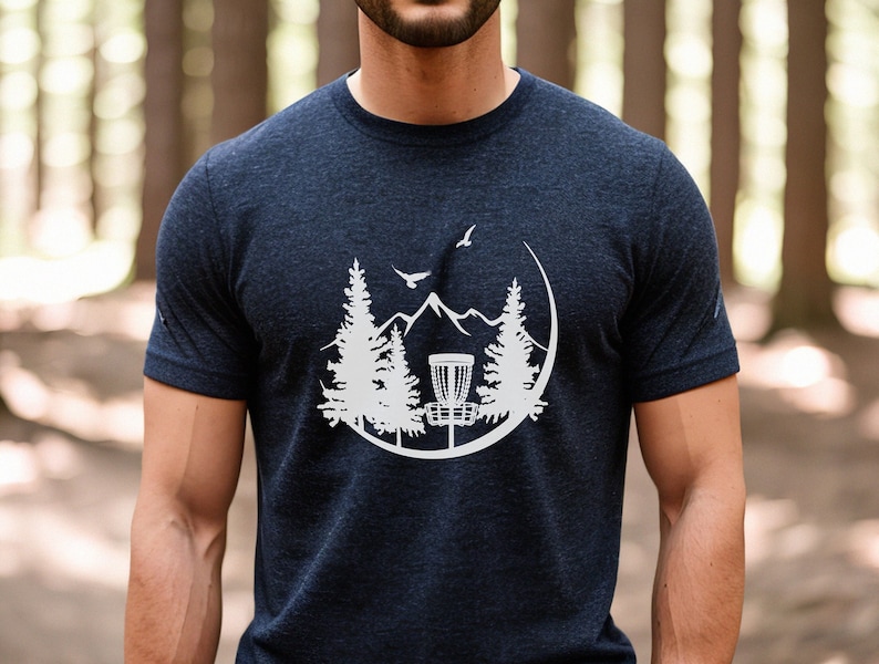 May include: A navy blue t-shirt featuring a white graphic of a disc golf basket, mountains, trees, and a bird flying overhead.