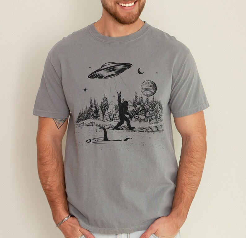 May include: A grey t-shirt featuring a black graphic print of a Bigfoot playing disc golf with a UFO flying overhead. The graphic also includes a Loch Ness Monster in the water.