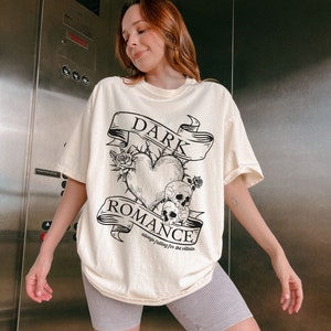 Op de afbeelding: Een cr&egrave;mekleurig T-shirt met een zwarte grafische print van een hart met doornen, schedels en een banner met de tekst "DARK ROMANCE always falling for the villain".