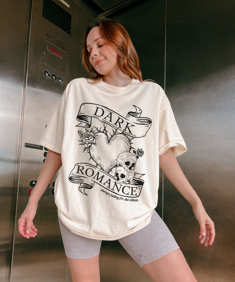Op de afbeelding: Een cr&egrave;mekleurig T-shirt met een zwarte grafische print van een hart met doornen, schedels en een banner met de tekst "DARK ROMANCE always falling for the villain".