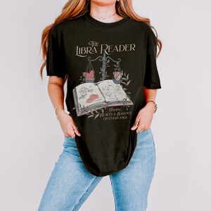 Pu&ograve; includere: Maglietta nera con un design grafico che presenta un libro, una bilancia e il testo "The Libra Reader: Finding Beauty & Balance on Every Page".