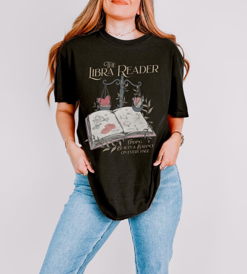 Pu&ograve; includere: Maglietta nera con un design grafico che presenta un libro, una bilancia e il testo "The Libra Reader: Finding Beauty & Balance on Every Page".