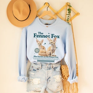 May include: Light blue sweatshirt with a graphic of two fennec foxes. The text "The Fennec Fox" is printed in dark blue above the foxes. The text "World's Smallest Fox" is in a circle below the foxes. The text "Also known as the Desert Fox" is printed below the circle. The text "Suspiciously Cute" is printed below the desert fox text.