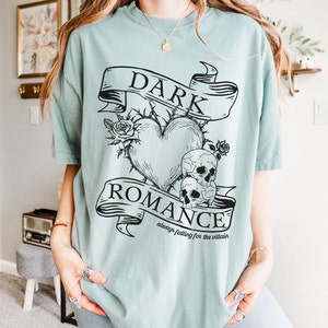 Op de afbeelding: Een lichtblauw t-shirt met een zwart grafisch ontwerp met een hart met doornen, twee schedels en een banner met de tekst "DARK ROMANCE always falling for the villain".