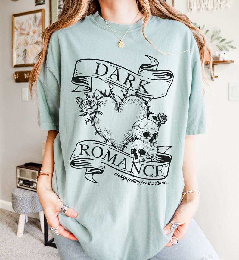Op de afbeelding: Een lichtblauw t-shirt met een zwart grafisch ontwerp met een hart met doornen, twee schedels en een banner met de tekst "DARK ROMANCE always falling for the villain".