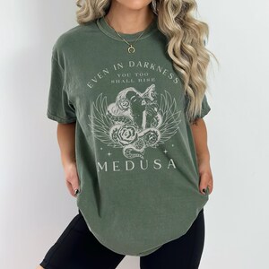 May include: Olive green t-shirt featuring a graphic of Medusa with the text "EVEN IN DARKNESS YOU TOO SHALL RISE MEDUSA". The design includes a snake, wings, and roses. The shirt is a relaxed fit.