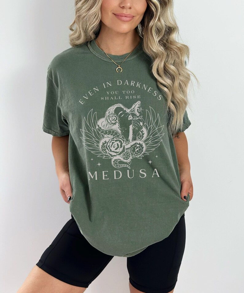 May include: Olive green t-shirt featuring a graphic of Medusa with the text "EVEN IN DARKNESS YOU TOO SHALL RISE MEDUSA". The design includes a snake, wings, and roses. The shirt is a relaxed fit.