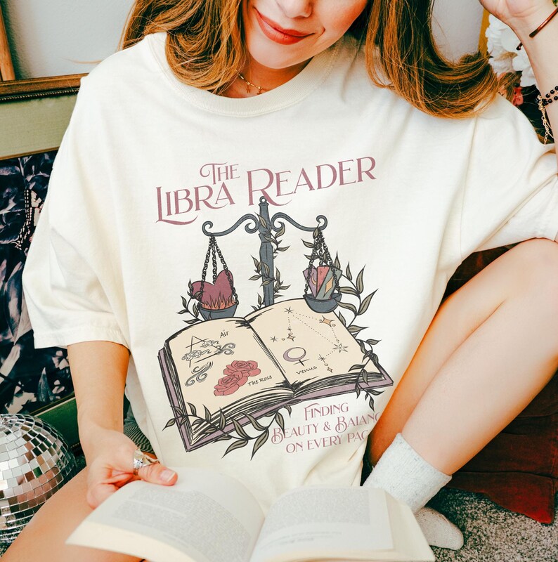 Pu&ograve; includere: Una maglietta bianca con un design grafico di un libro, una bilancia e viti. Il testo "The Libra Reader" &egrave; stampato sulla maglietta. Il testo "Finding Beauty & Balance on Every Page" &egrave; anche stampato sulla maglietta.