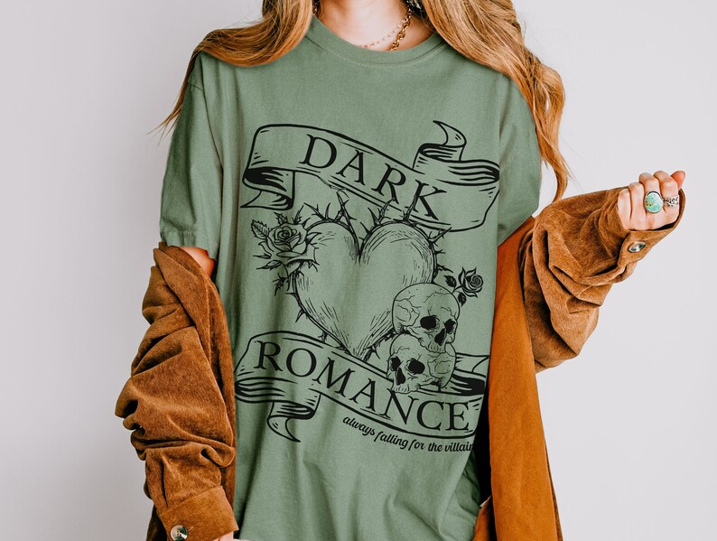 Op de afbeelding: Groen T-shirt met een zwart grafisch ontwerp van een hart met doornen, twee schedels en een banner met de tekst "DARK ROMANCE always falling for the villain".