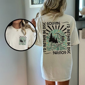 May include: A white t-shirt with a graphic print on the back. The graphic features a sunset, trees, a disc golf basket, and the text "I'd rather be disc golfing".