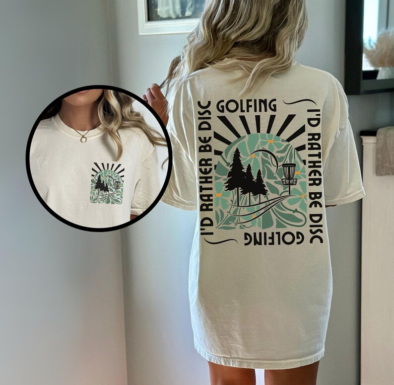 May include: A white t-shirt with a graphic print on the back. The graphic features a sunset, trees, a disc golf basket, and the text "I'd rather be disc golfing".