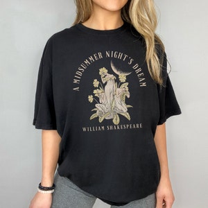May include: Black t-shirt featuring a graphic print of a moon, flowers, and three figures. The text "A Midsummer Night's Dream" is in a circle around the graphic. The text "William Shakespeare" is below the graphic.