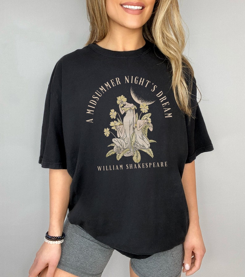May include: Black t-shirt featuring a graphic print of a moon, flowers, and three figures. The text "A Midsummer Night's Dream" is in a circle around the graphic. The text "William Shakespeare" is below the graphic.